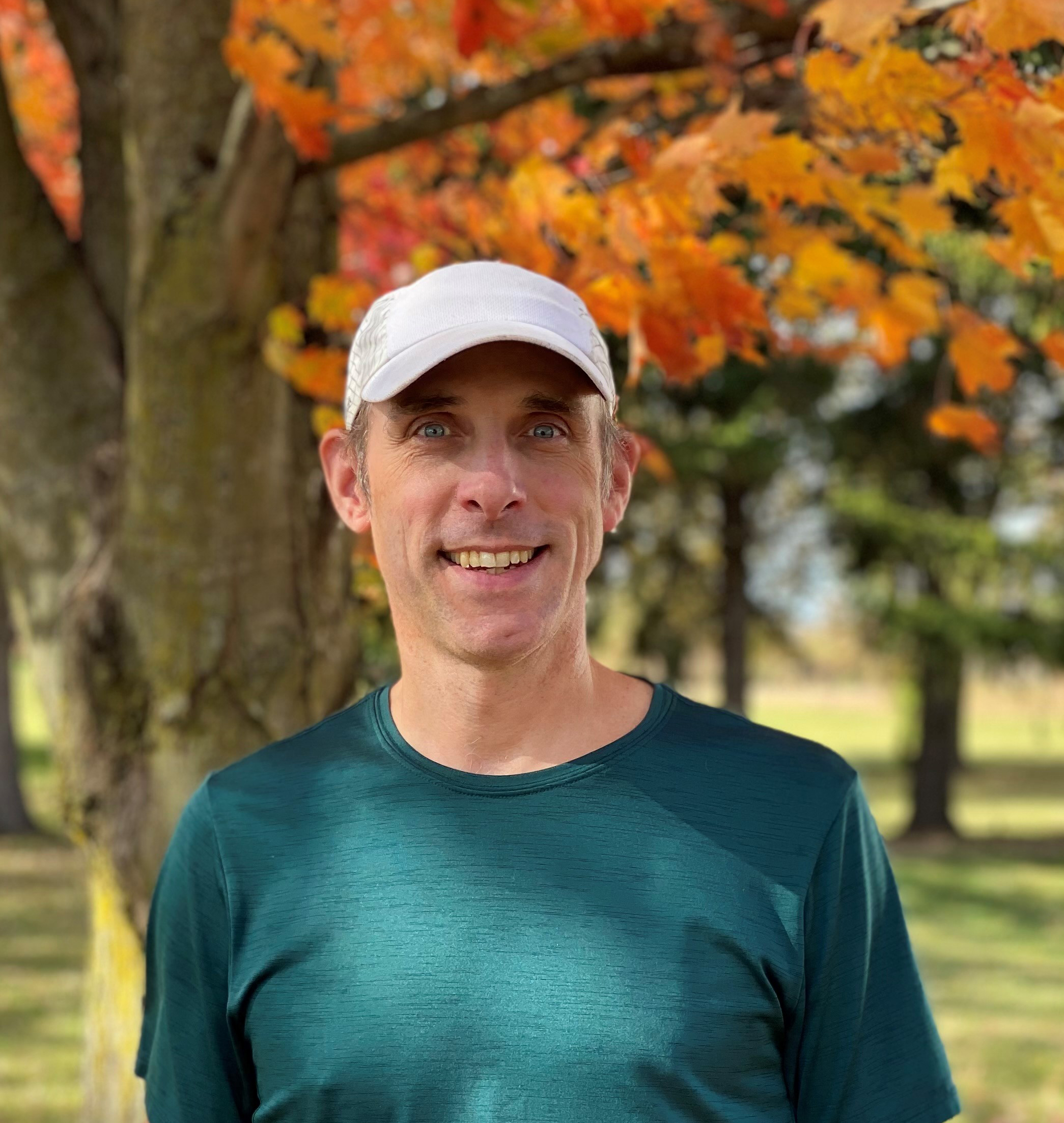 Outdoor portrait of Steve wearing a green shirt and a light-colored cap, standing in front of a tree with orange and yellow fall leaves.